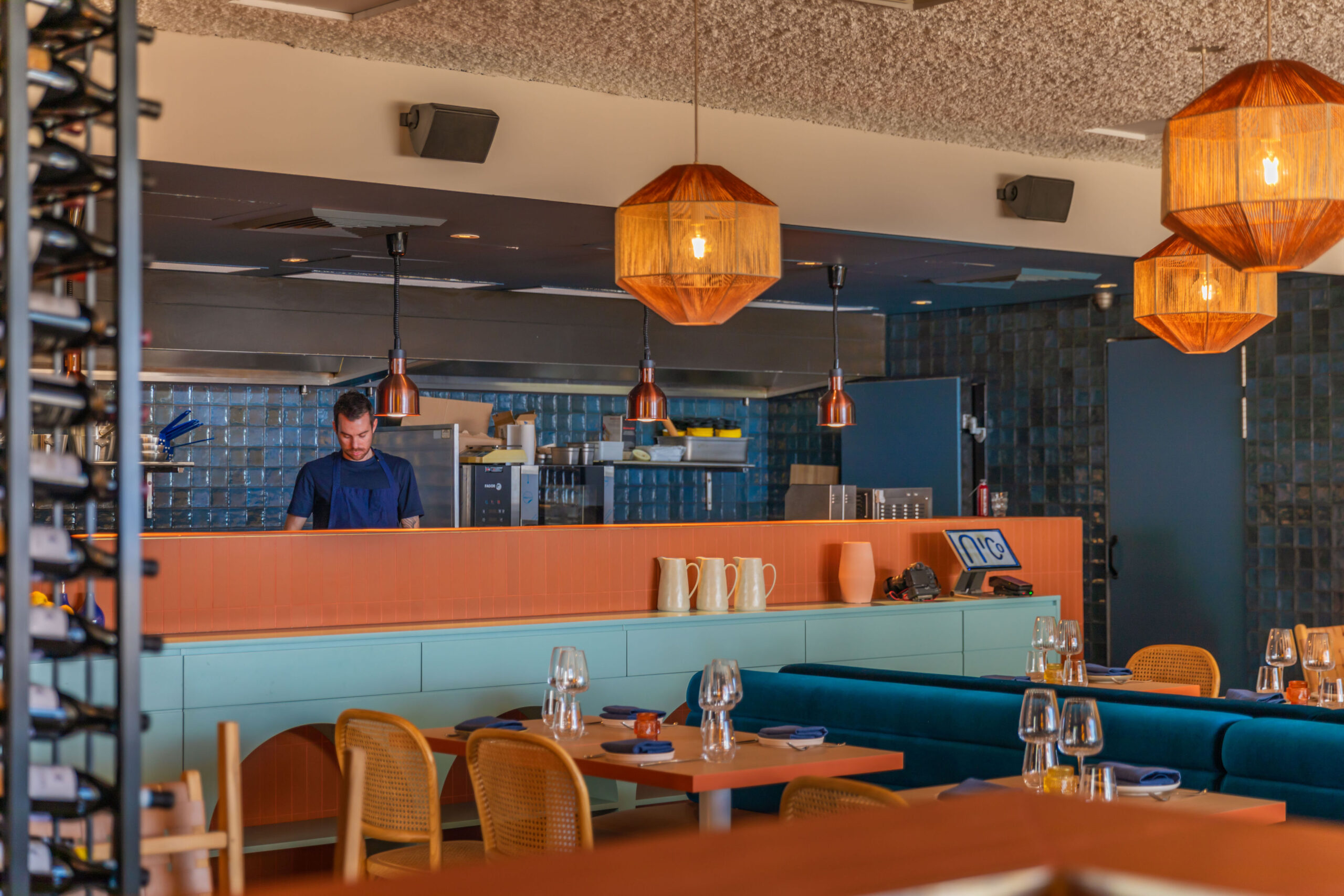 Vibrant restaurant interior with warm lighting, blue ceramic subway tiles on the walls, wooden furniture, and a mix of terracotta and teal decor elements.