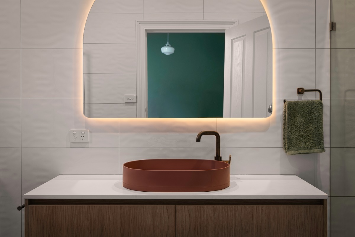 Bathroom vanity with a rust vessel sink, black faucet, arched backlit mirror, and white tiles with dark contrasting grout.