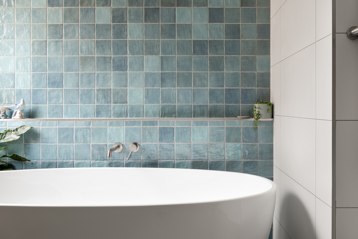 Freestanding white bathtub against a blue square-tiled wall with light gray grout, featuring wall-mounted silver faucets and a small potted plant on a narrow shelf.
