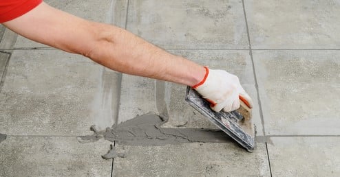 Worker in gloves applying gray grout to textured floor tiles using a grout float.